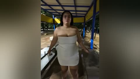 Video of a plus-size woman with medium skin tone, wearing a strapless white mini dress, standing under a yellow and blue canopy in a park.