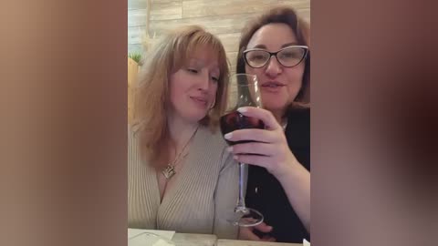 Media: Video of two women, one with light hair in a V-neck top, the other with glasses and brown hair in a black top, toasting with wine glasses in a cozy, beige-tiled room.