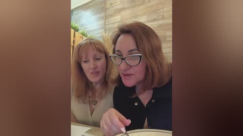 Media: Video of two middle-aged women with light skin, one with shoulder-length blonde hair, the other with shoulder-length brown hair. They are sitting close, smiling, and holding hands over a table, with a beige wall and green plant in the background.