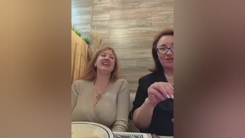 Media: Video of two women in a restaurant: one with shoulder-length blonde hair in a beige top, smiling, and the other with glasses, dark hair, in a black top, laughing.