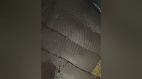 Video of a dimly lit, cluttered room with a textured, corrugated metal ceiling and exposed pipes. A yellow tarp is partially visible in the top right corner, adding a splash of color. The room appears disorganized, with various objects and shadows creating a chaotic atmosphere.