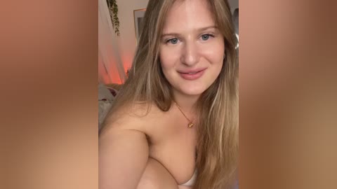 Video of a smiling, fair-skinned woman with long blonde hair, wearing a strapless top, sitting in a softly lit room. Background includes a bed with white sheets and a green plant.
