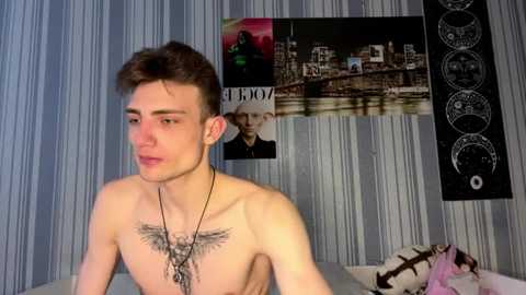 Video of a shirtless, slender, young man with short brown hair and a detailed tattoo on his chest, standing in a room with striped wallpaper, a poster, and a black tapestry with white circular designs.