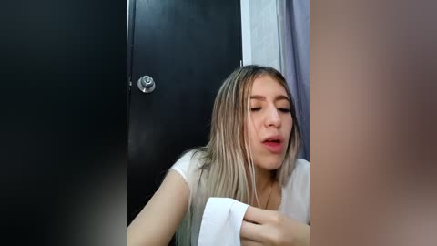 Media: Video of a young woman with fair skin, blonde hair, and light makeup, holding a white towel in a bathroom with a dark door and silver handle.