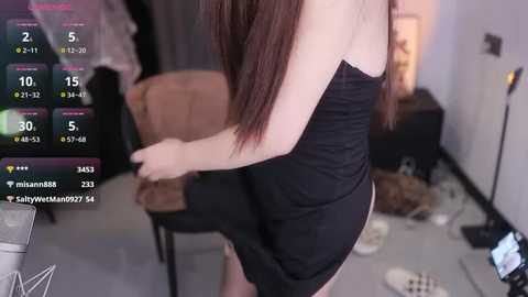 Video of a young, slender Asian woman with long, straight brown hair, wearing a tight, black strapless dress, standing in a cluttered room with a TV screen displaying social media metrics.