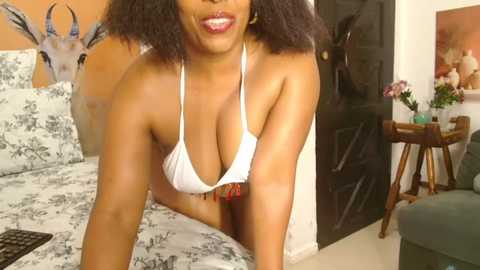 Video of a smiling Black woman with curly hair in a white bikini top, leaning forward on a bed with floral sheets. Background features a wooden table, deer head, and floral artwork.