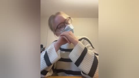 Video of a woman with glasses, wearing a white and black striped sweater, and a face mask, standing in a beige room with a white wall and a light fixture.
