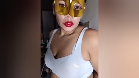 Video of a light-skinned woman with a medium build and large breasts, wearing a gold Venetian mask and a light blue sports bra, indoors with a blurred background.