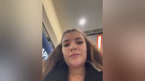 Video of a young woman with light skin, brown hair, and full lips, wearing a black jacket, standing in a brightly lit indoor space with a blurred background.