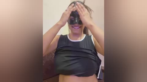 Media: Video of a woman in a dark room, wearing a black mask, black tank top, and white shorts, hands over eyes. Background is blurred.
