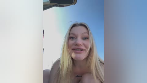 Video of a smiling young woman with long blonde hair, fair skin, and a tattoo on her chest, standing in front of a light-colored wall with a bright blue sky background.