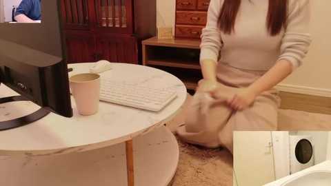 Video of a person in a beige sweater and pants kneeling on a carpeted floor, working on a circular white table with a computer, keyboard, and cup. Background features wooden furniture and a doorway.
