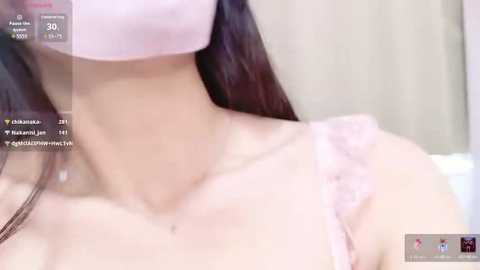 Media: Video of a light-skinned woman with long black hair, wearing a pink face mask, revealing a delicate neck and shoulder with a sheer, lace-trimmed top.
