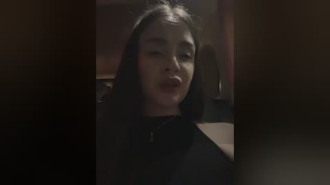 Media: A dimly-lit video captures a young woman with long, straight black hair, wearing a black top, slightly squinting and pursing her lips. The background is dark and indistinct, suggesting an indoor setting with low light.