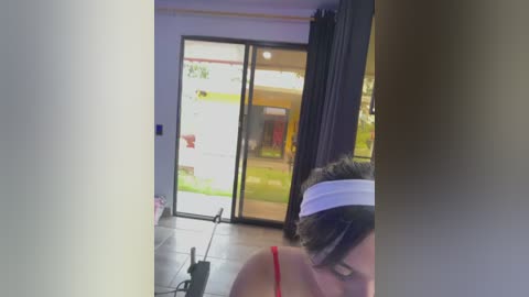 Media: A video of a woman in a red tank top and white headband, taken from a side angle, standing in front of a sliding glass door with a green lawn outside.