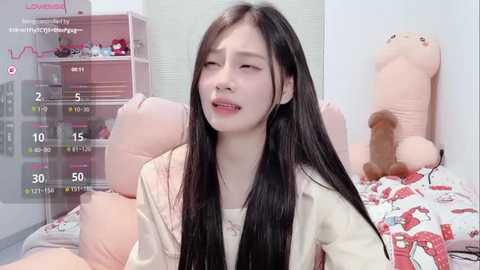 Media: A video of a young Asian woman with long black hair, wearing a cream-colored robe, lying on a bed with a stuffed bear. The room is pink-themed, with a clock displaying 5:10 PM.