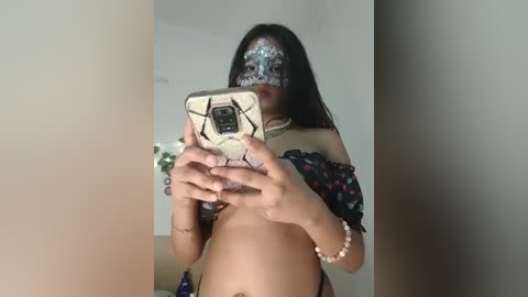 Media: Video of a young Asian woman with long black hair, wearing a silver face mask and a black floral off-shoulder top, taking a selfie in a dimly lit room.