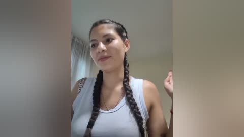 Video of a young woman with long, braided hair, wearing a white tank top, standing indoors near a window with white curtains.