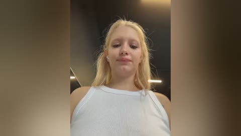 Media: A video of a blonde Caucasian woman with fair skin, wearing a white ribbed tank top, standing in a dimly lit room with blurred background.