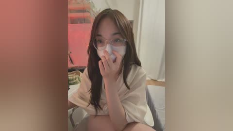 A video of an Asian woman with long dark hair, wearing glasses, and a white shirt, holding a finger to her lips, sitting on a bed. The background shows a cluttered room with a pinkish hue.