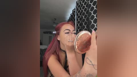 Video of a young woman with pink hair and multiple piercings, including a septum ring, applying makeup in a bedroom with a black and white geometric-patterned curtain.