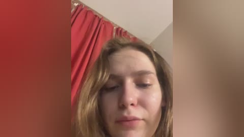 A video of a young woman with light skin and straight, shoulder-length blonde hair, partially obscured by a red curtain in a dimly lit room.