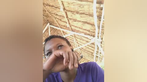 Video of a young woman with medium brown skin, dark hair, and a broad smile, wearing a purple shirt. She rests her chin on her hand, surrounded by a thatched-roof hut.