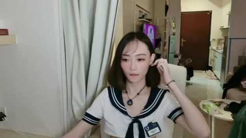 Media: Video of an Asian woman with straight black hair, wearing a Japanese schoolgirl uniform, sitting in a cluttered, dimly lit room, adjusting her hair.