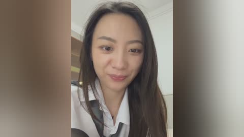 A close-up video of a young Asian woman with long, straight black hair, fair skin, and a light smile. She's wearing a white shirt and black tie. The background is a blurred, neutral-toned room.