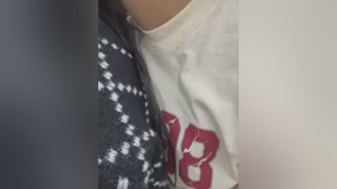 A close-up video of a person's torso, showing a white t-shirt with a red \"18\" graphic and a gray sweater with a white diamond pattern. The image is slightly blurry, and the background is plain and neutral-colored.