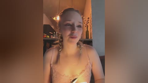 Media: Video of a young woman with light skin and blonde braided hair, wearing a light pink lace camisole, smiling, set indoors with warm lighting, kitchen background.