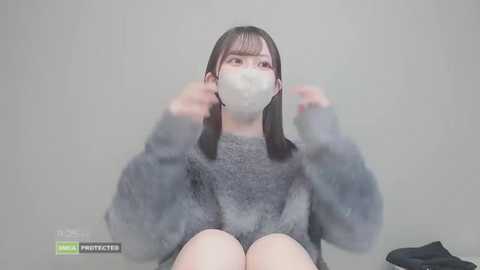 Video of a young Asian woman with straight black hair, wearing a grey sweater, white mask, and grey pants, sitting against a plain light grey wall.