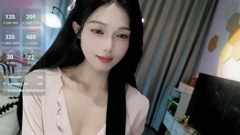 Video of an East Asian woman with long black hair, fair skin, and a slender figure, wearing a light pink blouse. Background includes a desk with a computer monitor, a lamp, and a bookshelf.