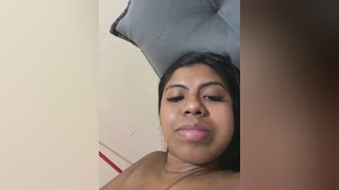 Video of a young Black woman with medium brown skin, straight black hair, and full lips, lying on a bed with a grey pillow, smiling slightly.