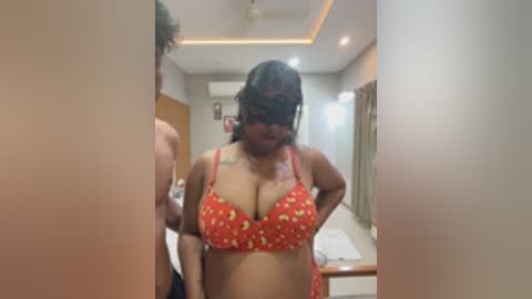 Media: Video of a curvy, dark-skinned woman with large breasts in a red floral bra, standing in a modern, beige-tiled bathroom with a mirror, air conditioner, and partially visible person.