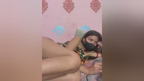 Media: A video shows a young woman with dark hair, wearing a black mask and colorful dress, lying on a bed with blue and red floral wallpaper in the background.