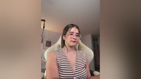 Video of a young woman with light skin and straight dark hair, wearing glasses and a brown and white striped crop top, sitting in a white gaming chair in a dimly lit room.