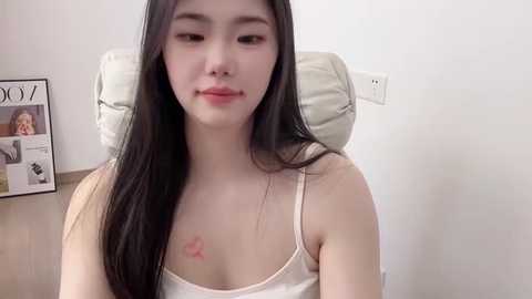 Video of a young Asian woman with long black hair, fair skin, wearing a white tank top, sitting in a chair against a white wall.