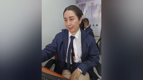 Media: Video of a smiling, light-skinned woman with brown hair in a ponytail, wearing a navy jacket, white shirt, and black tie, seated at a desk in an office. Background shows another person and a partially visible poster.