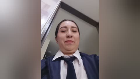 Media: Video of a woman with light skin and dark hair, dressed in a navy blue security uniform, standing in a beige-walled hallway, looking straight at the camera with a neutral expression.