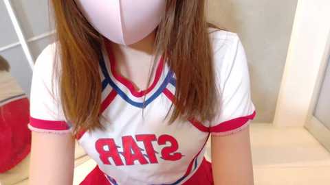 Video of a young woman with long, straight brown hair, wearing a white cheerleader outfit with red trim, \"RATS\" on the chest, and a white mask, indoors with a beige wall and red gym bag in the background.