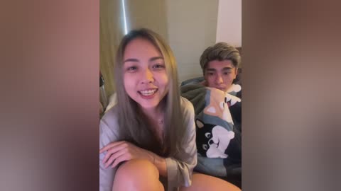 Video of a young Asian woman with light brown hair, smiling, wearing a grey hoodie, sitting next to a boy in a dark shirt with cartoon characters, both indoors.