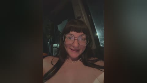 Media: Video of a smiling woman with long brown hair and glasses, wearing a white dress, inside a dimly lit room with a slanted ceiling.