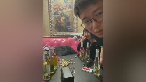 A video captures two Asian individuals with glasses, sitting at a table in a dimly lit, vibrant room with a framed mural on the wall. The table is cluttered with bottles, snacks, and a phone charger.