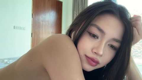 A video of a young Asian woman with long black hair, light skin, and full lips, topless, lying on her side, resting her head on her hand, in a modern room with a wooden door and large windows.