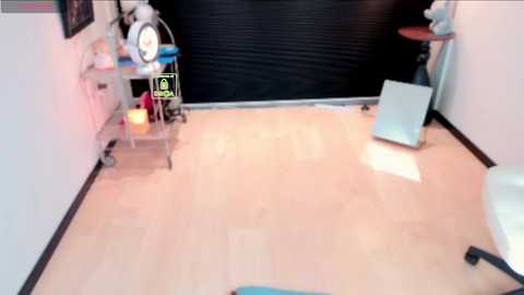 A video of a modern medical office with a light wood floor, a black rolling cabinet, and a white exam chair. A large, closed black door and various medical equipment are visible.