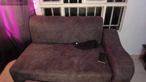 A video of a worn-out, brown, L-shaped sofa in a dimly lit room with purple lighting. A black cat sleeps on the sofa, and a smartphone rests on the armrest. The background includes a white-framed window and a dark curtain.