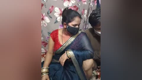 Video of a South Asian woman in traditional attire with a red blouse, blue sari, and gold bangles, sitting in a room with floral-patterned wallpaper, wearing a mask, and being tended to by a man in a brown shirt.