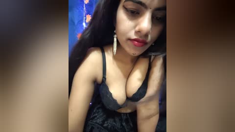Media: Video of a young South Asian woman with long black hair, medium skin tone, wearing a black lace bra revealing ample cleavage, and red lipstick. Background includes a blue and orange patterned wall.