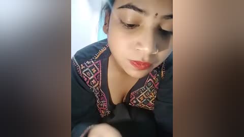 Media: Video of a South Asian woman with fair skin, wearing a traditional black blouse with colorful geometric patterns, focused on applying makeup, eyes closed.